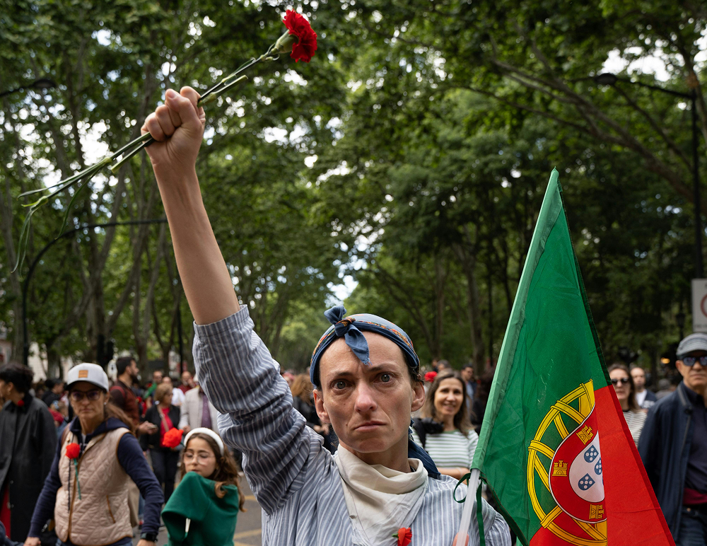 Pessoa segura cravos no alto e a bandeira de Portugal como símbolo da revolução dos cravos.